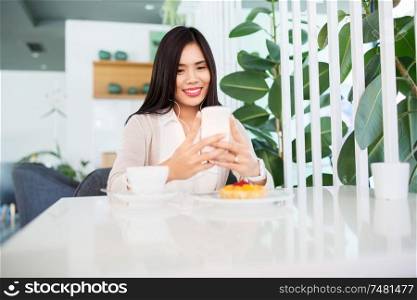 technology and people concept - happy asian woman with smartphone, earphones and cake listening to music at cafe or coffee shop. asian woman with smartphone and earphones at cafe