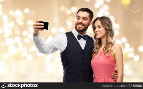 technology and people concept - happy couple in party clothes taking selfie by smartphone over beige background with festive lights. happy couple taking selfie by smartphone