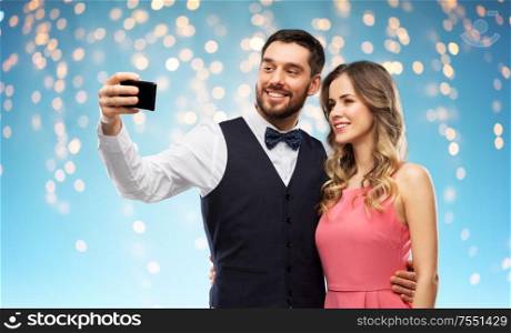 technology and people concept - happy couple in party clothes taking selfie by smartphone over holiday lights on blue background. happy couple taking selfie by smartphone