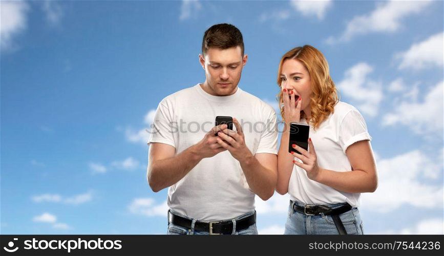 technology and people concept - happy couple in white t-shirts with smartphones over blue sky and clouds background. happy couple in white t-shirts with smartphones