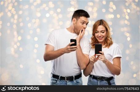 technology and people concept - happy couple in white t-shirts with smartphones over holidays lights background. happy couple in white t-shirts with smartphones