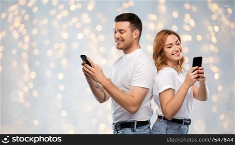 technology and people concept - happy couple in white t-shirts with smartphones over holidays lights background. happy couple in white t-shirts with smartphones