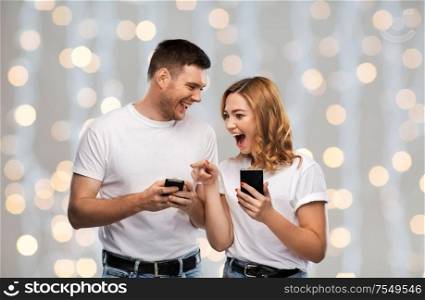 technology and people concept - happy couple in white t-shirts with smartphones over festive lights background. happy couple in white t-shirts with smartphones