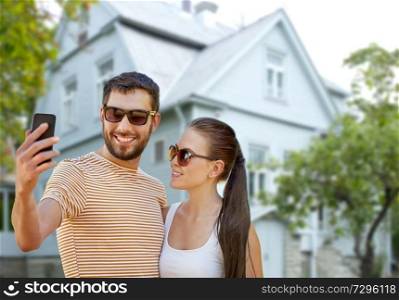 technology and people concept - happy couple taking selfie by smartphone in summer over house background. couple taking selfie by smartphone over house