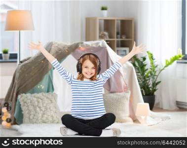 technology and people concept - happy girl with headphones listening to music at home at home over kids room and tepee background. girl with headphones listening to music at home