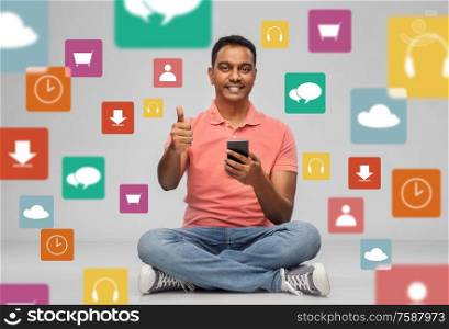 technology and people concept - happy indian man with smartphone showing thumbs up over mobile app icons on grey background. happy indian man with smartphone showing thumbs up