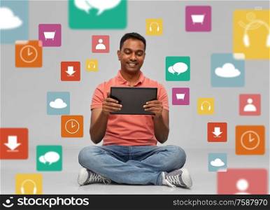 technology and people concept - happy indian man with tablet computer sitting on floor over mobile app icons on grey background. happy indian man with tablet pc over app icons