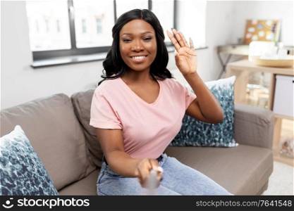 technology and people concept - happy smiling african american woman with selfie stick waving hand and taking picture on sofa at home. happy african american woman taking selfie at home