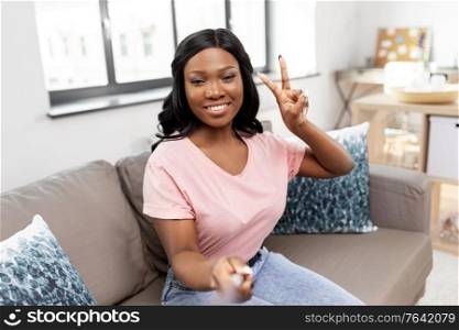 technology and people concept - happy smiling african american woman with selfie stick showing peace hand sign taking picture on sofa at home. happy african american woman taking selfie at home