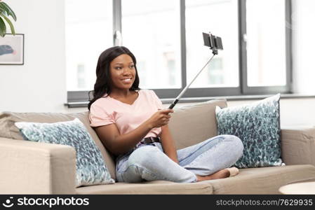 technology and people concept - happy smiling african american woman with smartphone and selfie stick taking picture sitting on sofa at home. happy african american woman taking selfie at home