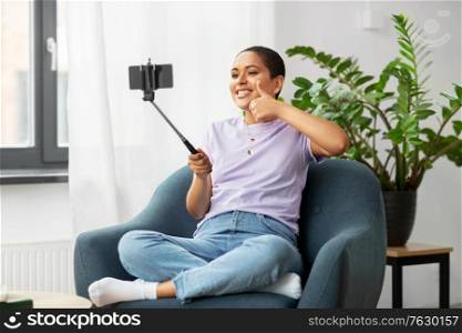 technology and people concept - happy smiling african american woman with smartphone and selfie stick taking picture sitting in chair at home. happy african american woman taking selfie at home