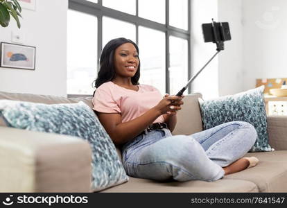 technology and people concept - happy smiling african american woman with smartphone and selfie stick taking picture sitting on sofa at home. happy african american woman taking selfie at home