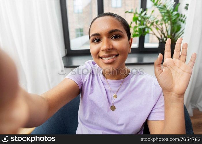 technology and people concept - happy smiling african american woman with taking selfie sitting in chair and waving hand at home. happy african american woman taking selfie at home