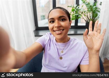 technology and people concept - happy smiling african american woman with taking selfie sitting in chair and waving hand at home. happy african american woman taking selfie at home