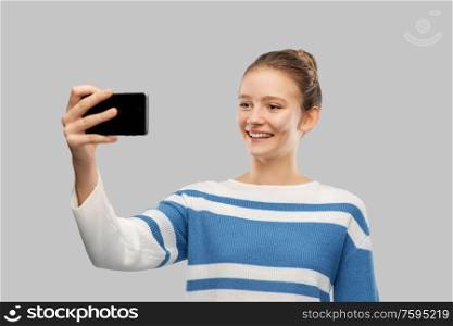 technology and people concept - happy smiling teenage girl in pullover taking selfie with smartphone over grey background. smiling teenage girl taking selfie with smartphone