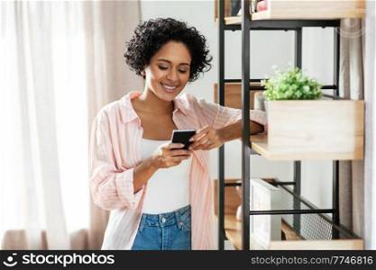 technology and people concept - happy smiling woman with smartphone standing at shelf at home. smiling woman with smartphone at home