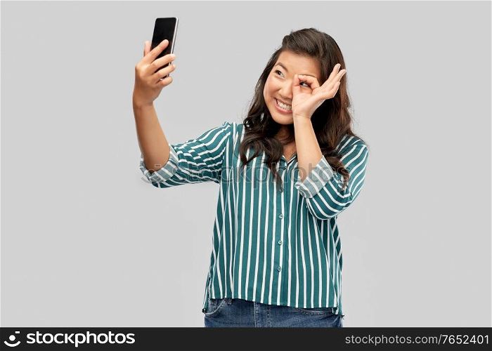 technology and people concept - happy smiling young asian woman taking selfie by smartphone over grey background. smiling asian woman taking selfie by smartphone
