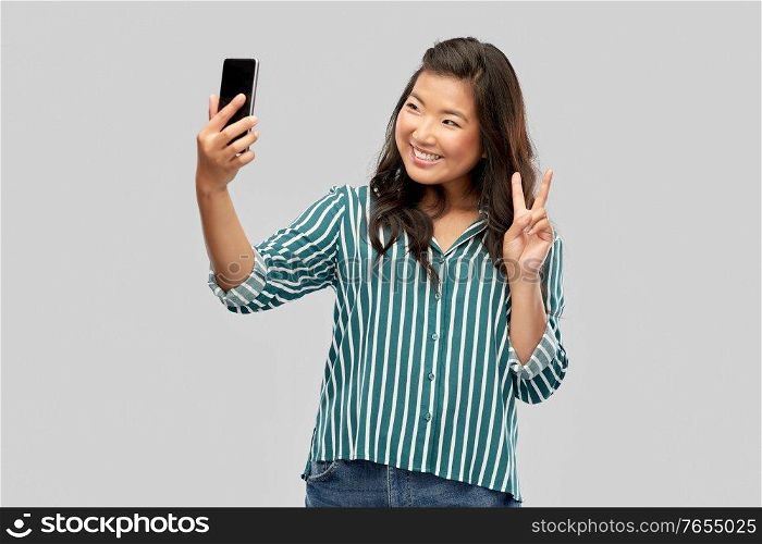 technology and people concept - happy smiling young asian woman taking selfie by smartphone showing peace hand sign over grey background. smiling asian woman taking selfie by smartphone