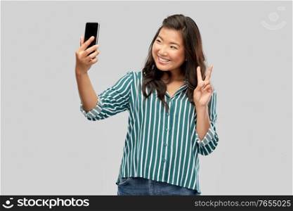 technology and people concept - happy smiling young asian woman taking selfie by smartphone showing peace hand sign over grey background. smiling asian woman taking selfie by smartphone