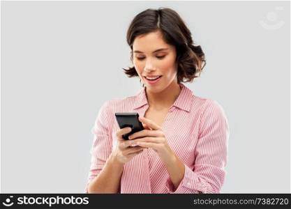 technology and people concept - happy smiling young woman in striped shirt using smartphone over grey background. young woman in striped shirt using smartphone