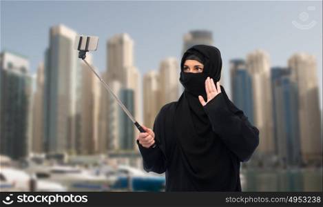 technology and people concept - muslim woman in hijab taking picture with smartphone selfie stick over dubai city street background. muslim woman in hijab taking selfie by smartphone