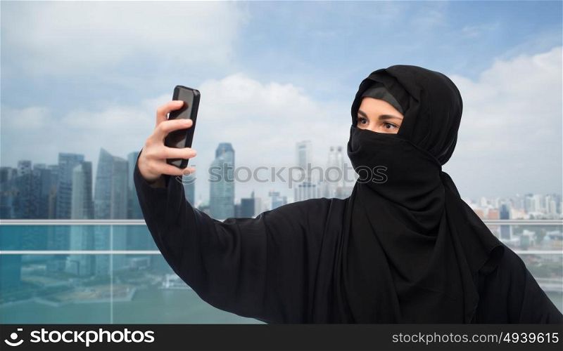 technology and people concept - muslim woman in hijab taking selfie with smartphone over singapore city skyscrapers background. muslim woman in hijab taking selfie by smartphone