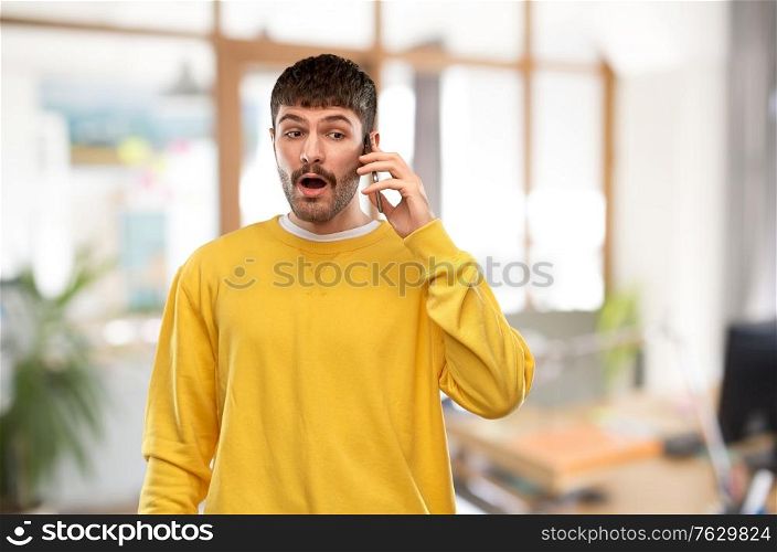 technology and people concept - shocked young man in yellow sweatshirt calling on smartphone over office background. shocked young man calling on smartphone