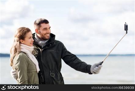 technology and people concept - smiling couple taking picture by smartphone on selfie stick on beach in autumn. happy couple taking selfie on beach in autumn