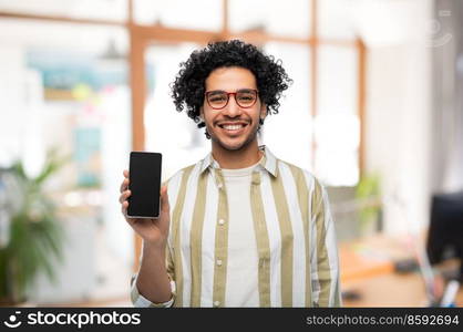 technology and people concept - smiling man in glasses showing smartphone with blank screen over office background. happy man in glasses showing smartphone at office