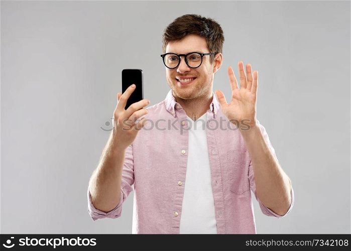 technology and people concept - smiling young man in glasses having video call on smartphone and waving hand over grey background. smiling man having video call on smartphone