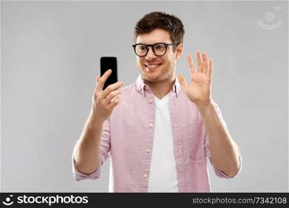 technology and people concept - smiling young man in glasses having video call on smartphone and waving hand over grey background. smiling man having video call on smartphone