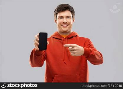 technology and people concept - smiling young man in red hoodie showing black smartphone with blank screen over grey background. smiling man in red hoodie showing black smartphone