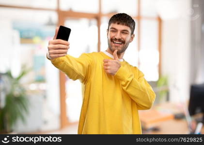 technology and people concept - smiling young man in yellow sweatshirt taking selfie with smartphone showing thumbs up over office background. man takes selfie with phone and shows thumbs up