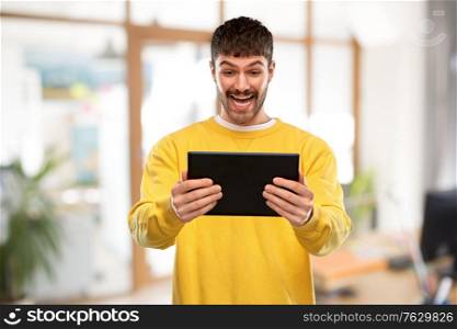 technology and people concept - smiling young man in yellow sweatshirt with tablet pc computer over office background. smiling young man with tablet pc computer