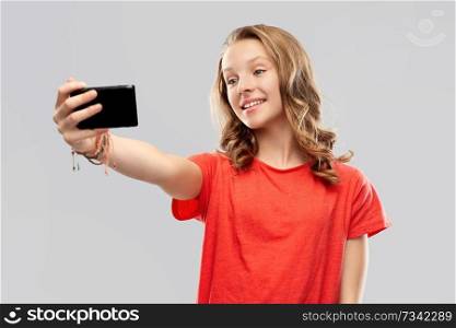 technology and people concept - smiling young woman or teenage girl in blank red t-shirt taking selfie by smartphone over grey background. smiling teenage girl taking selfie by smartphone