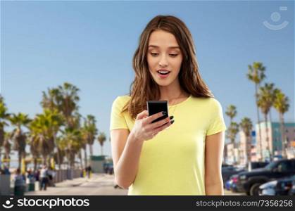 technology and people concept - smiling young woman or teenage girl in blank yellow t-shirt using smartphone over venice beach background in california. teenage girl using smartphone over venice beach