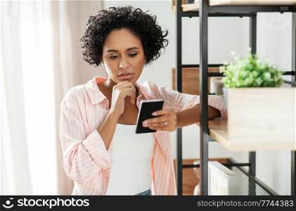 technology and people concept - woman with smartphone standing at shelf at home. woman with smartphone thinking at home