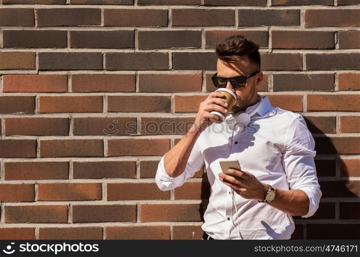 technology and people concept - young man in sunglasses with smartphone drinking coffee on city street. man with smartphone and coffee cup on city street