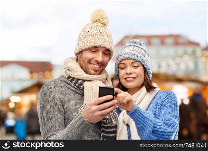technology and winter holidays concept - happy couple in knitted hats and scarves with smartphone over christmas market in old town of tallinn city background. happy couple with smartphone christmas market