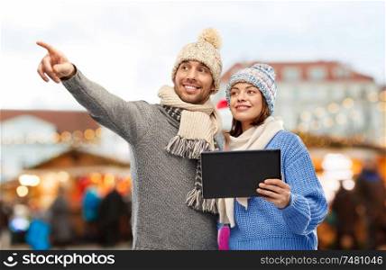 technology and winter holidays concept - happy couple in knitted hats and scarves with tablet computer over christmas market in old town of tallinn city background. couple with tablet computer at christmas market