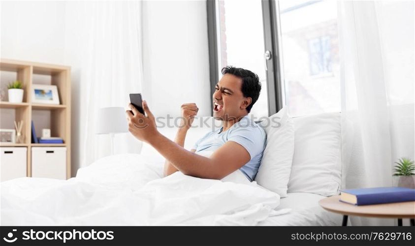 technology, bedtime and emotions concept - happy smiling indian man with smartphone lying in bed and celebrating success at home. happy indian man with smartphone in bed at home