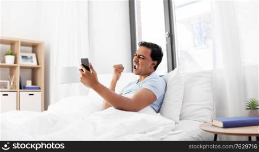 technology, bedtime and emotions concept - happy smiling indian man with smartphone lying in bed and celebrating success at home. happy indian man with smartphone in bed at home
