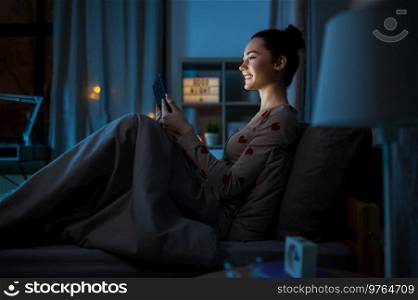 technology, bedtime and people concept - happy smiling teenage girl tablet pc computer sitting in bed at home at night. teenage girl with tablet pc in bed at night