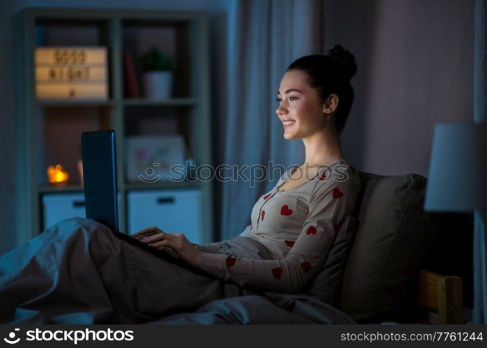 technology, bedtime and people concept - happy smiling teenage girl with laptop computer sitting in bed at home at night. teenage girl with laptop in bed at home at night