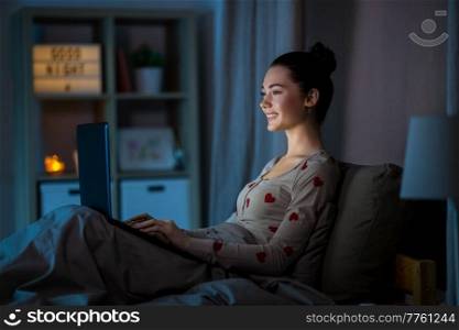 technology, bedtime and people concept - happy smiling teenage girl with laptop computer sitting in bed at home at night. teenage girl with laptop in bed at home at night