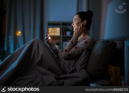 technology, bedtime and people concept - happy smiling teenage girl with smartphone and earphones sitting in bed at home at night. teenage girl with phone and earphones in bed at night