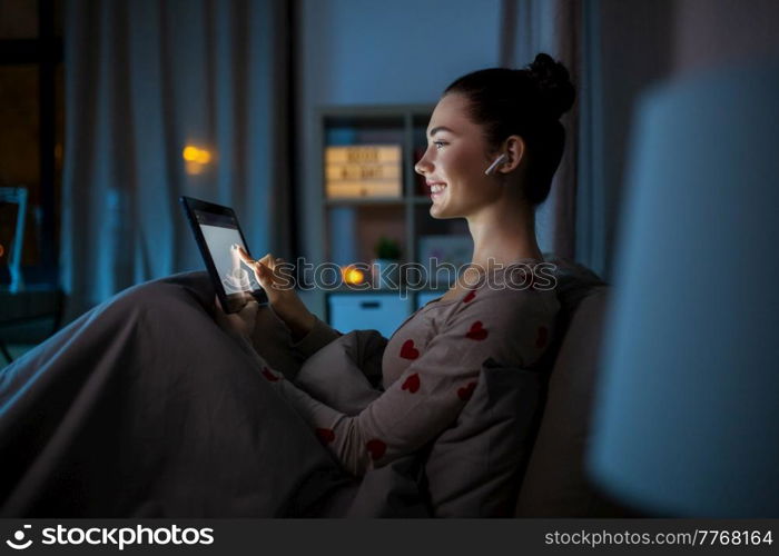 technology, bedtime and people concept - happy smiling teenage girl with tablet pc computer and earphones sitting in bed at home at night. teenage girl with tablet pc and earphones in bed