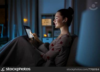 technology, bedtime and people concept - happy smiling teenage girl with tablet pc computer and earphones sitting in bed at home at night. teenage girl with tablet pc and earphones in bed