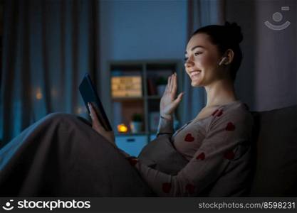 technology, bedtime and people concept - happy smiling teenage girl with tablet pc computer and earphones having video call sitting in bed at home at night and waving hand. teenage girl having video call on tablet pc in bed