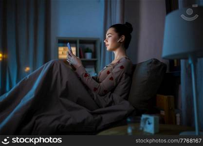 technology, bedtime and people concept - teenage girl with smartphone and earphones sitting in bed at home at night. teenage girl with phone and earphones in bed